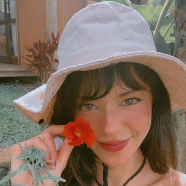 Milene Amorim sorrindo, de chapéu claro, segurando uma flor vermelha perto do rosto